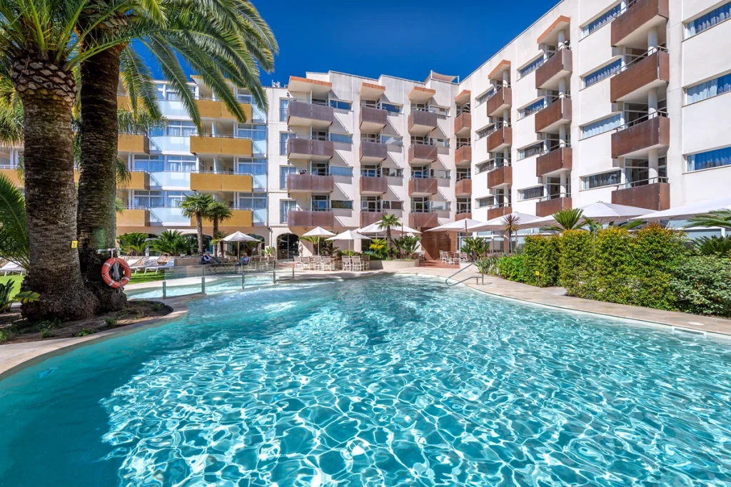 Piscina turquesa de hotel rodeada de terraza,palmeras y c&eacute;sped verde.