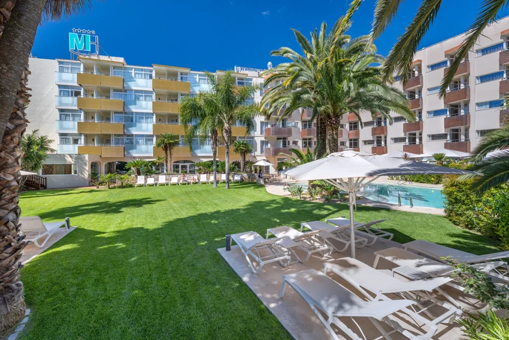 Jard&iacute;n de hotel con palmeras y tumbonas bajo un cielo despejado.