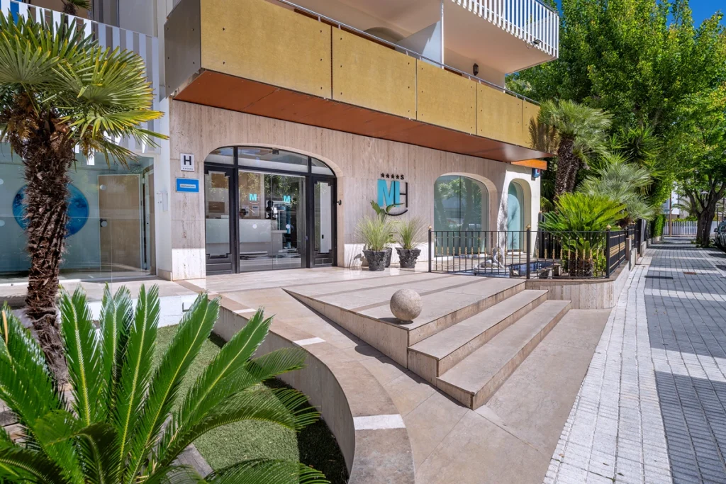 Entrada al Hotel con plantas y escalones, edificio moderno y fachada clara.