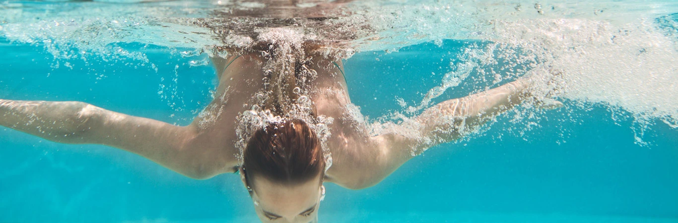 Persona nadando bajo el agua, brazos extendidos, burbujas y reflejos de sol.