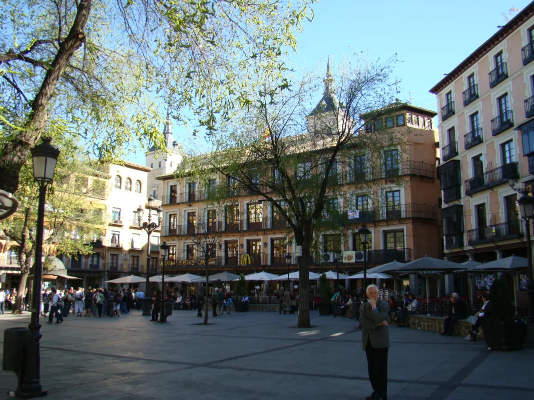 Plaza urbana con edificios cl&aacute;sicos, terrazas y gente disfrutando del sol.