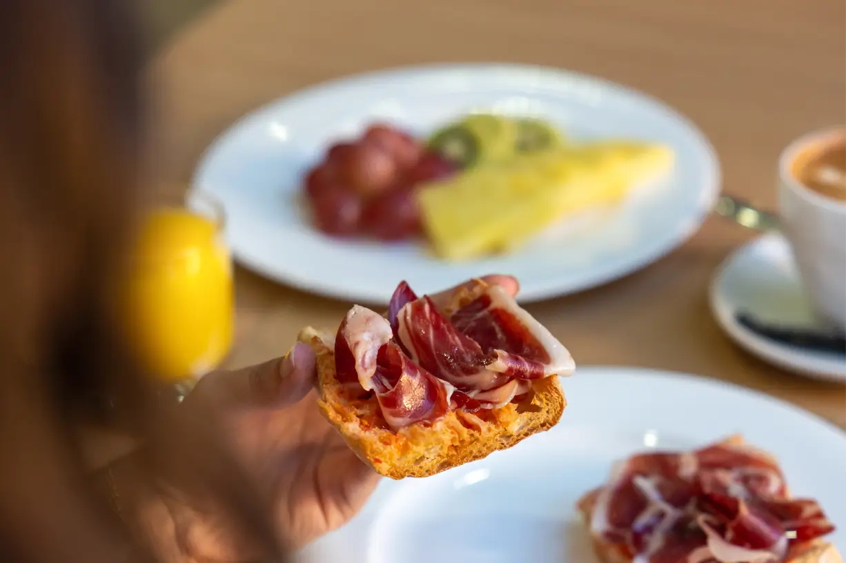 A person holding a slice of gourmet toast topped with ham, with a plate of more food in the background.