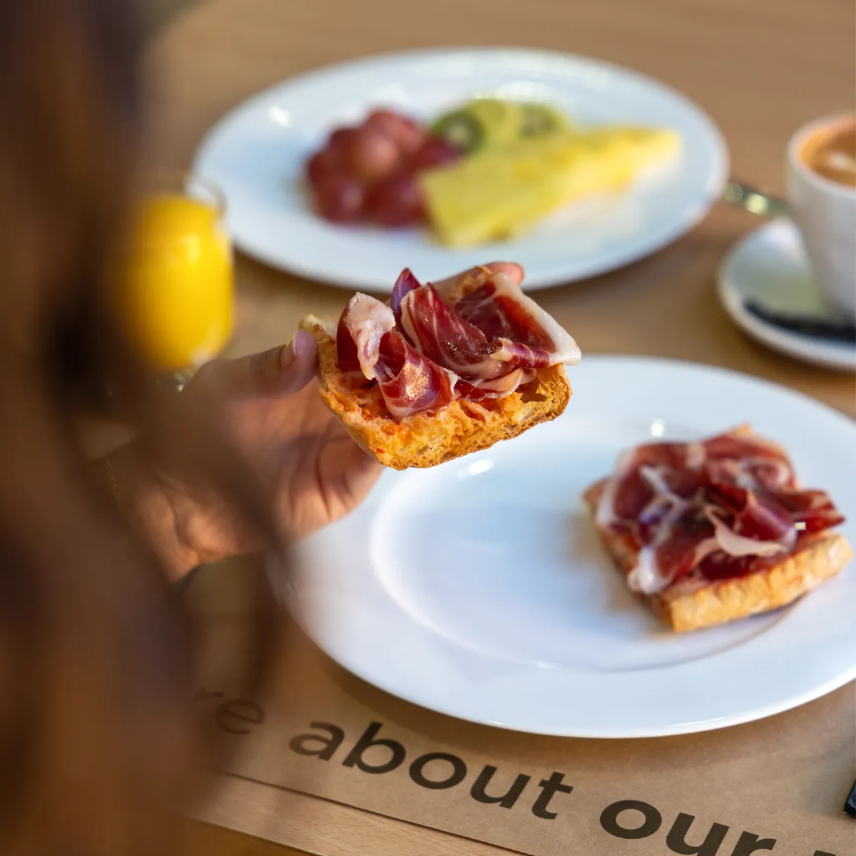 A person holding a slice of gourmet toast topped with ham, with a plate of more food in the background.