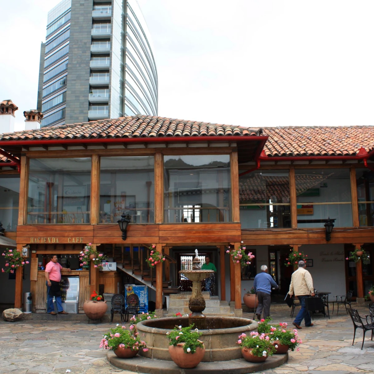 Fuente con flores y cafeter&iacute;a en patio, con edificio alto al fondo.