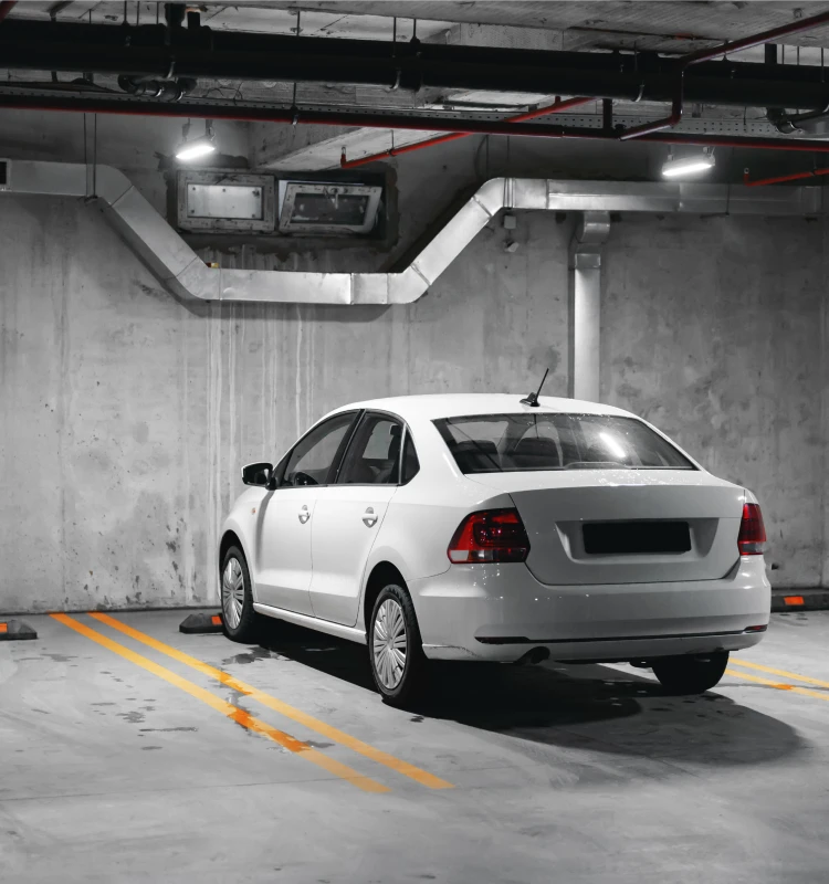 Auto blanco estacionado en un garaje subterr&aacute;neo, con marcas amarillas y cables arriba.