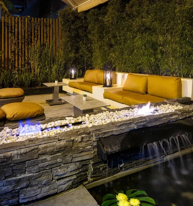 Patio nocturno con sof&aacute; amarillo, fogonera y pared de piedra con agua.