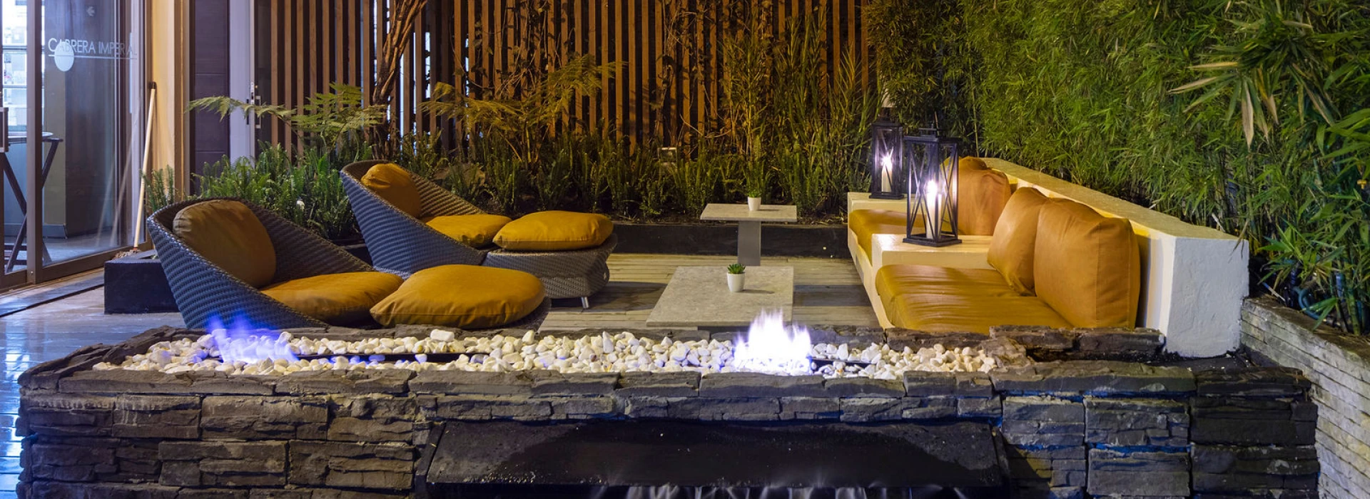 Jard&iacute;n interior con fogata, cojines amarillos y bamb&uacute; iluminado al atardecer