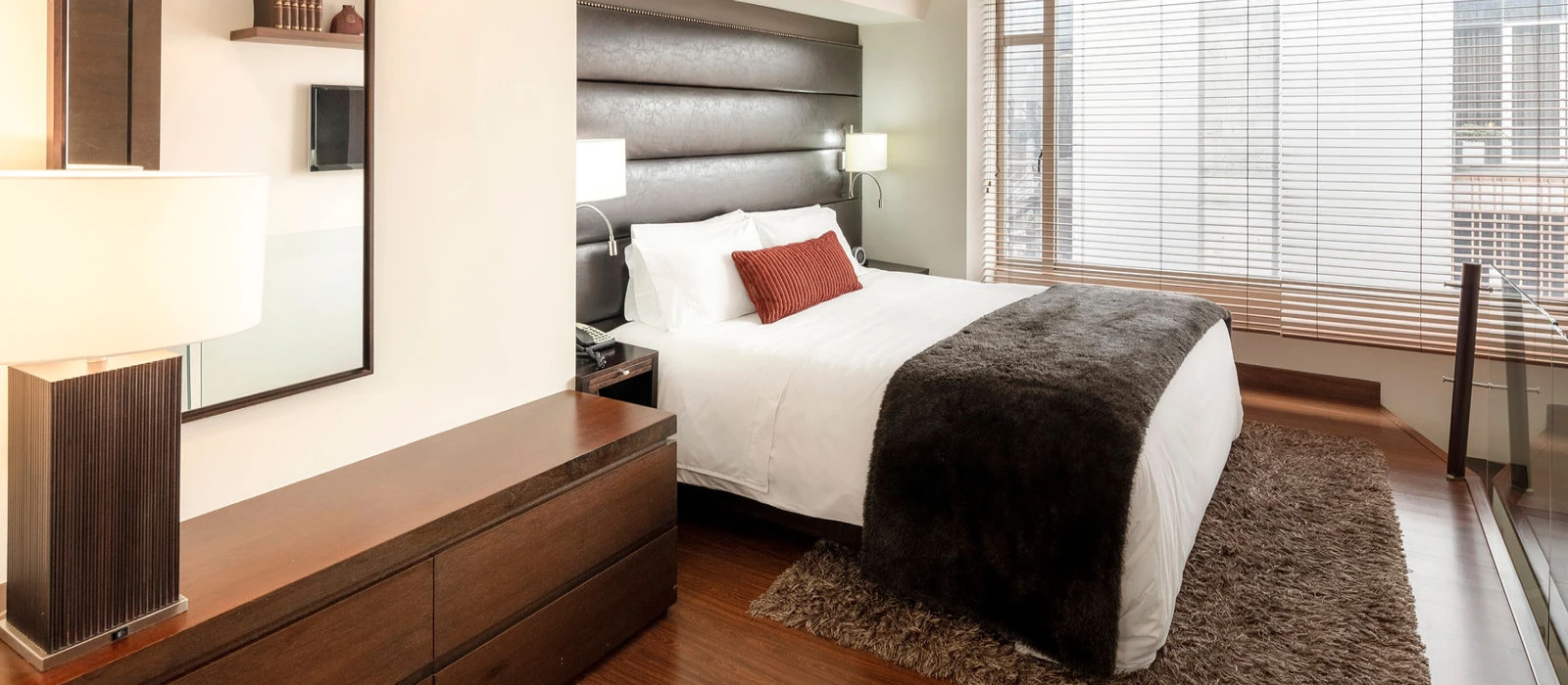 Dormitorio moderno con cama doble, manta gris y grandes ventanales con persianas