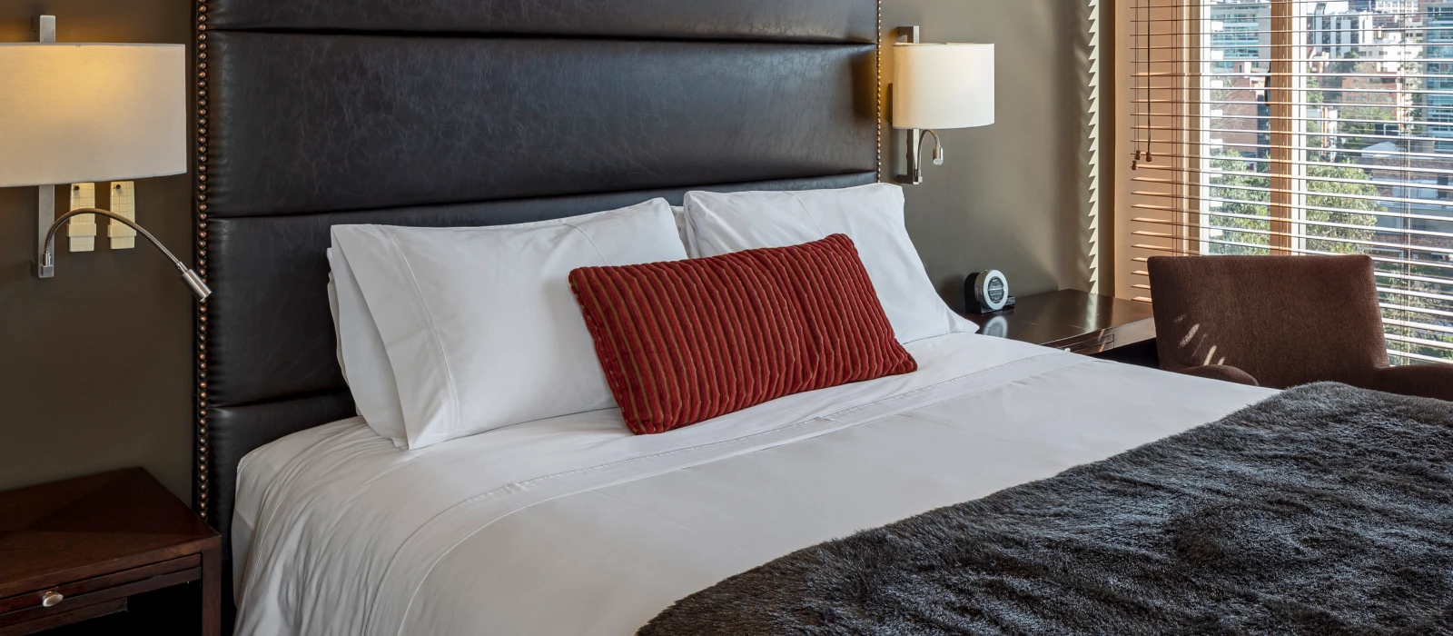 Cama con coj&iacute;n rojo y ropa de cama blanca, l&aacute;mparas y ventana con cortinas.