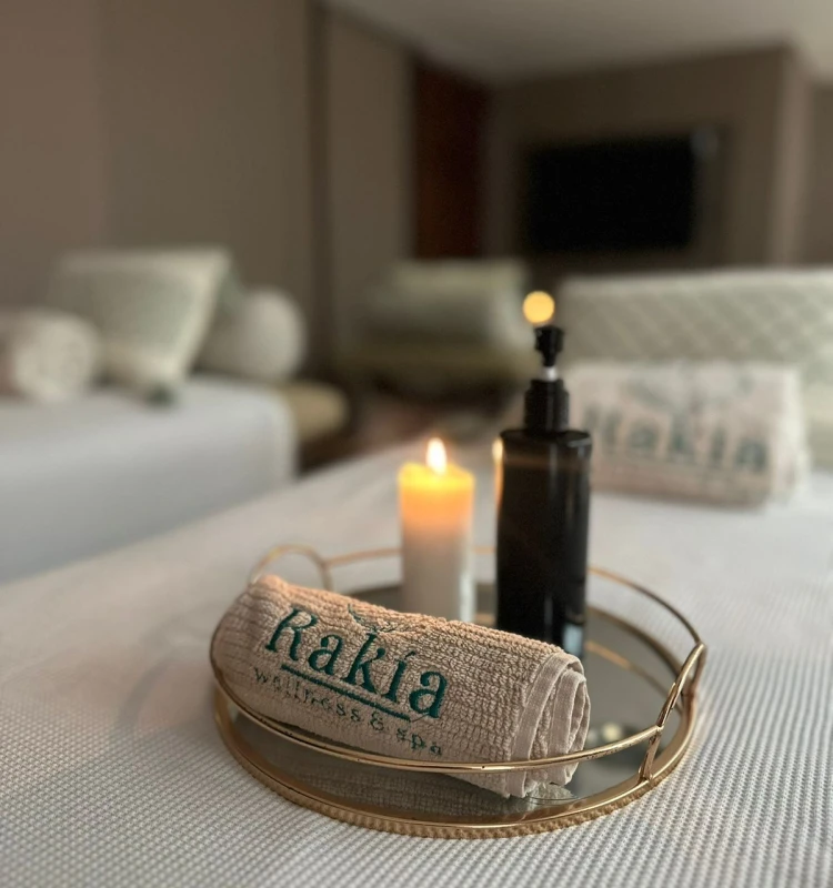 Toalla &ldquo;Rakiа&rdquo; en bandeja dorada con vela encendida y habitaci&oacute;n desenfocada