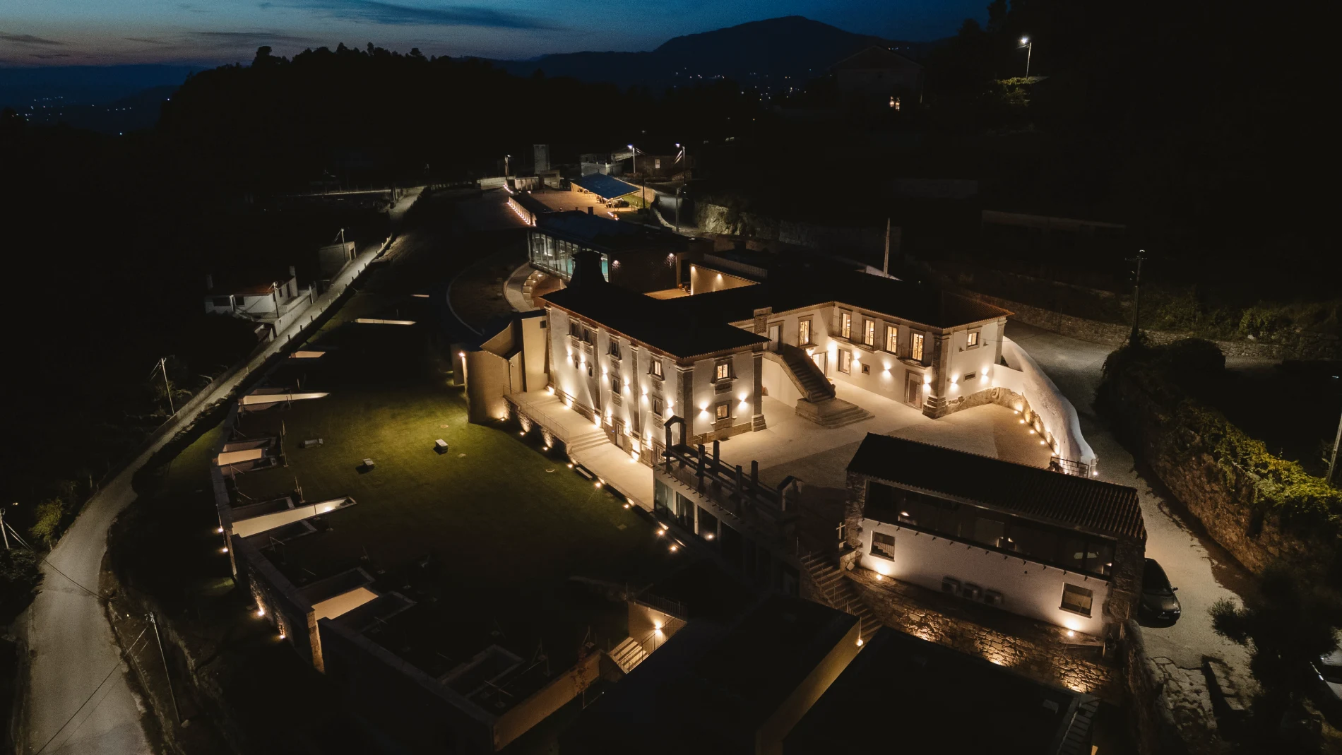 Vista a&eacute;rea de mans&atilde;o iluminada &agrave; noite, cercada por vegeta&ccedil;&atilde;o e montanhas.