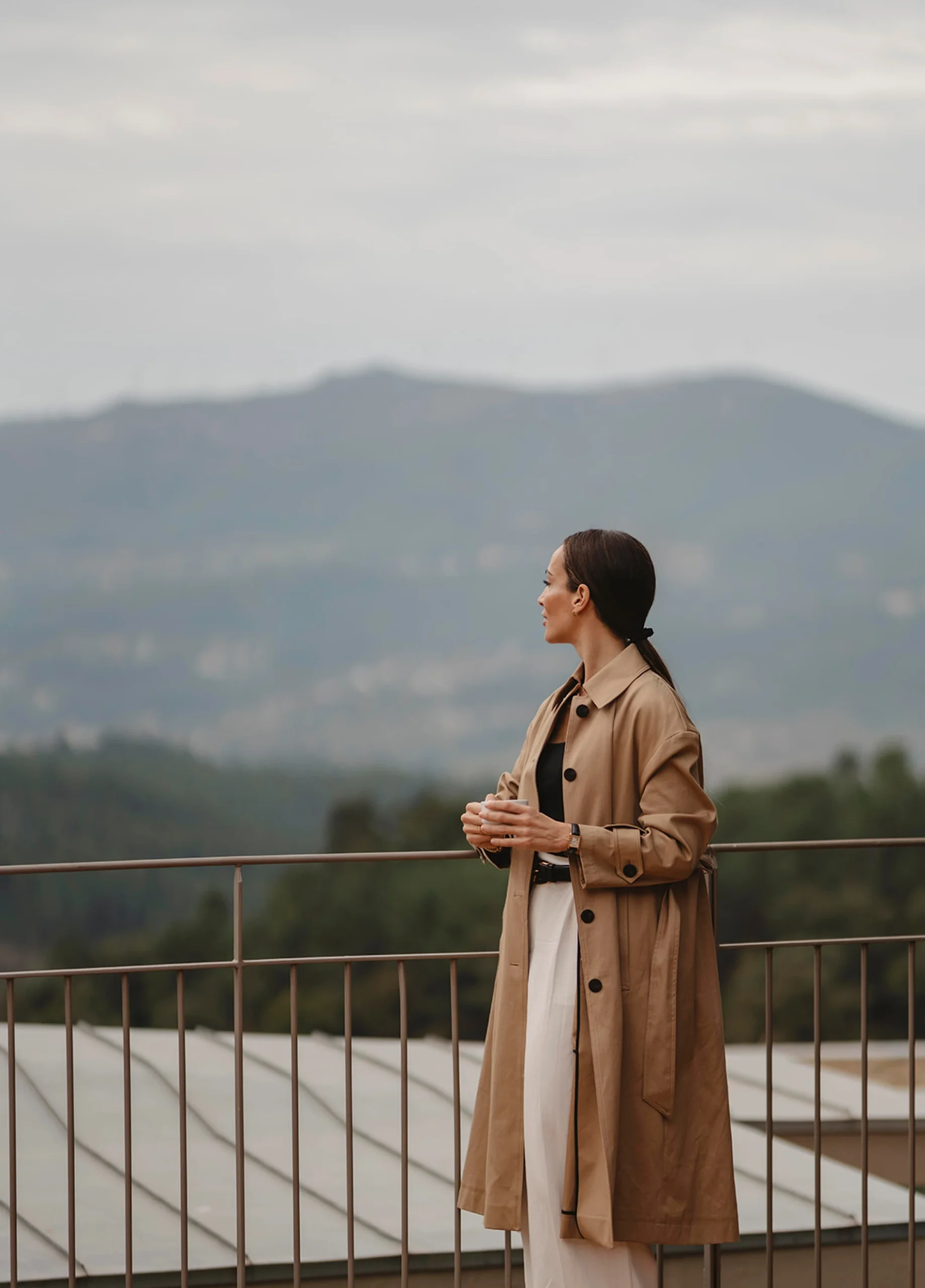 Mulher contemplando montanhas em um terra&ccedil;o, com casaco bege e x&iacute;cara na m&atilde;o.