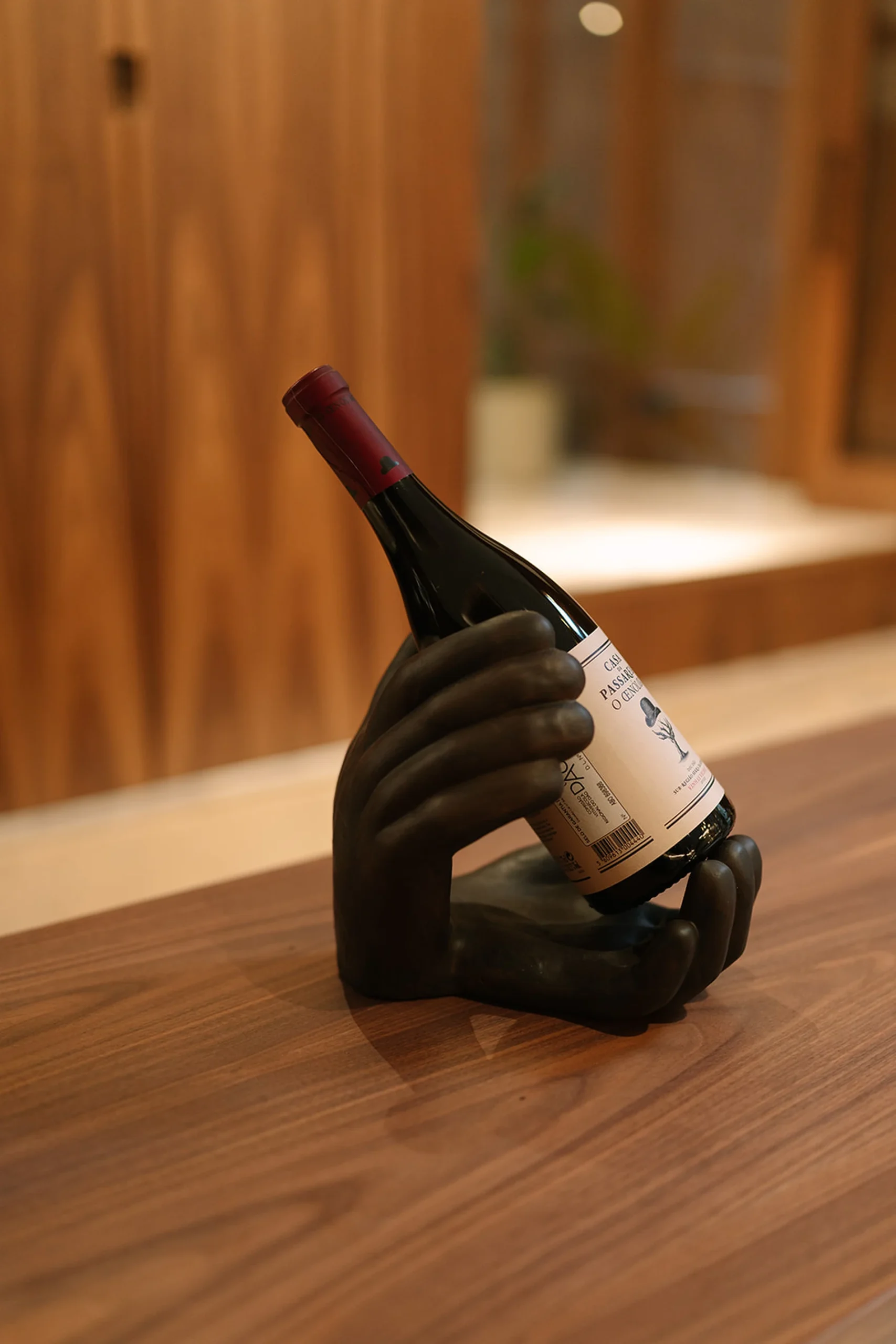 Garrafa de vinho em suporte de m&atilde;o escura sobre mesa de madeira.