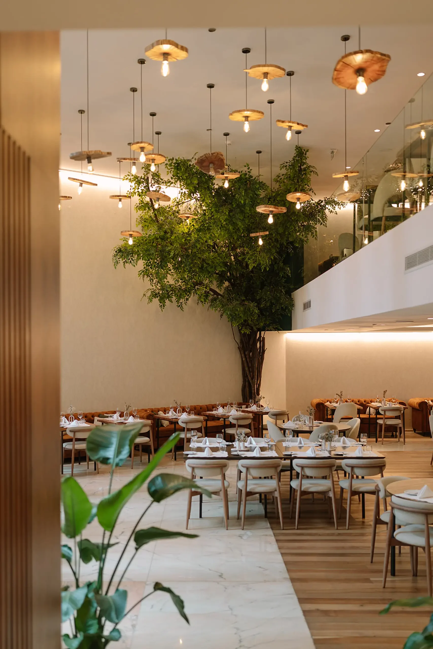 Restaurante elegante com mesas, plantas e lumin&aacute;rias pendentes.