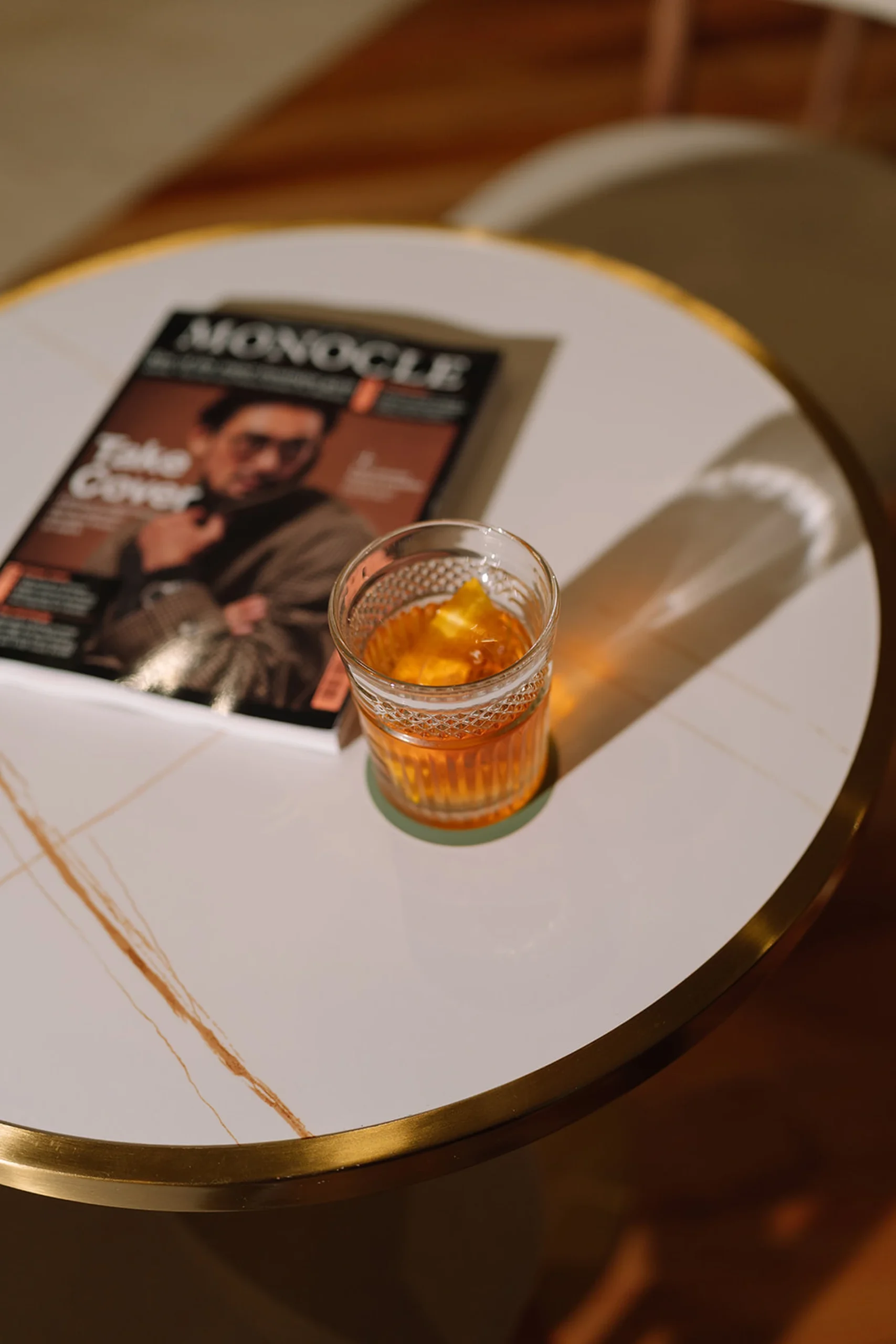 Copo de bebida e revista em uma mesa branca com borda dourada.