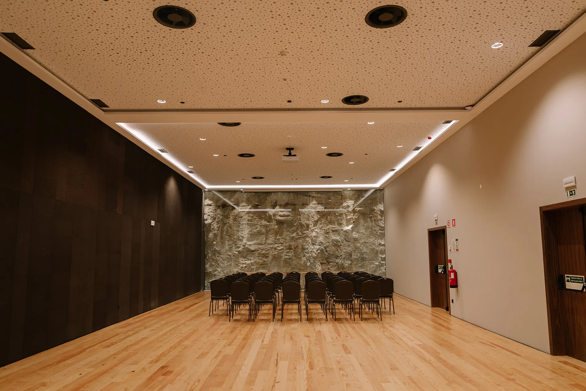 Sala de confer&ecirc;ncias vazia, com cadeiras e parede de pedra ao fundo.