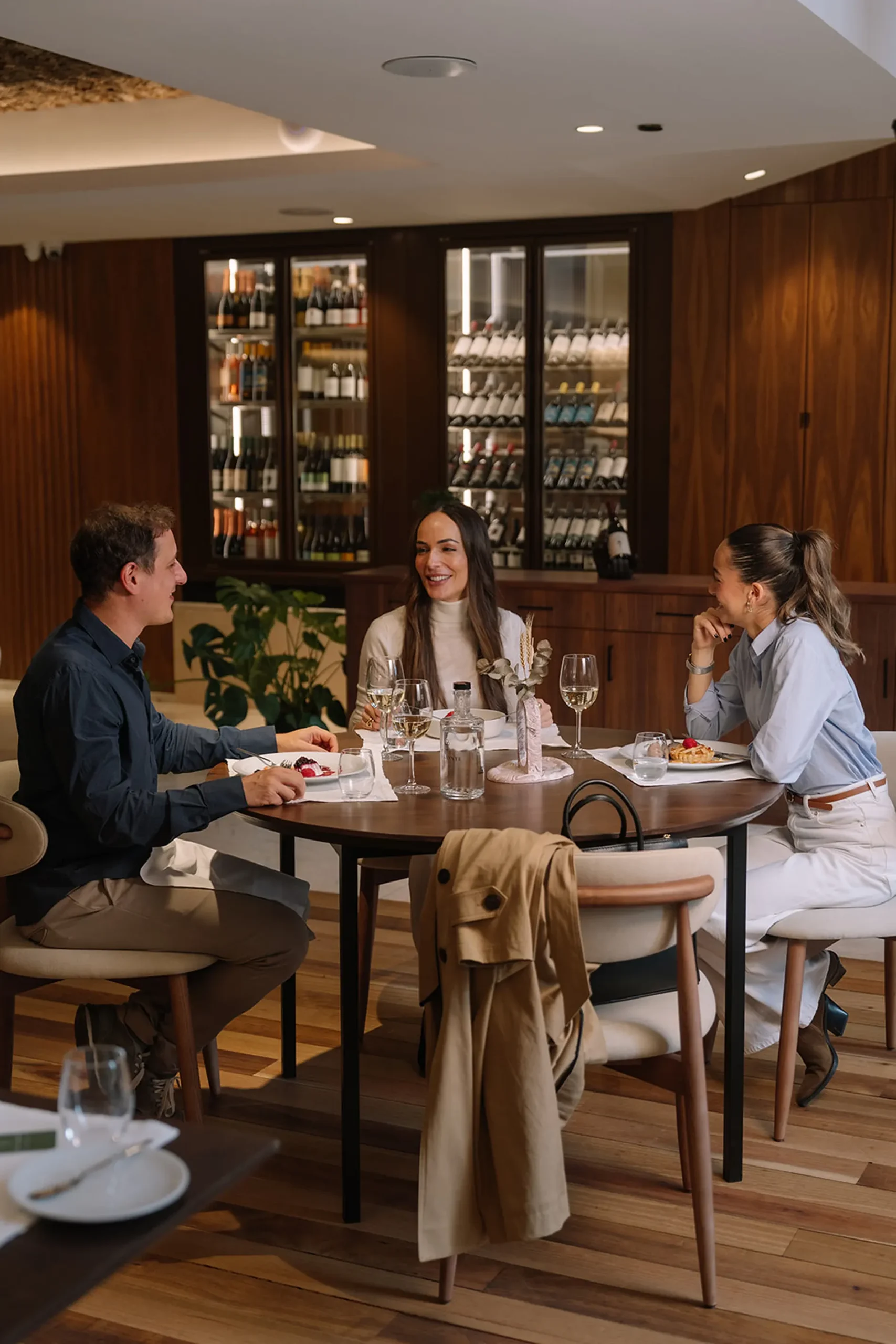 Tr&ecirc;s pessoas conversando em um restaurante elegante com adega ao fundo.