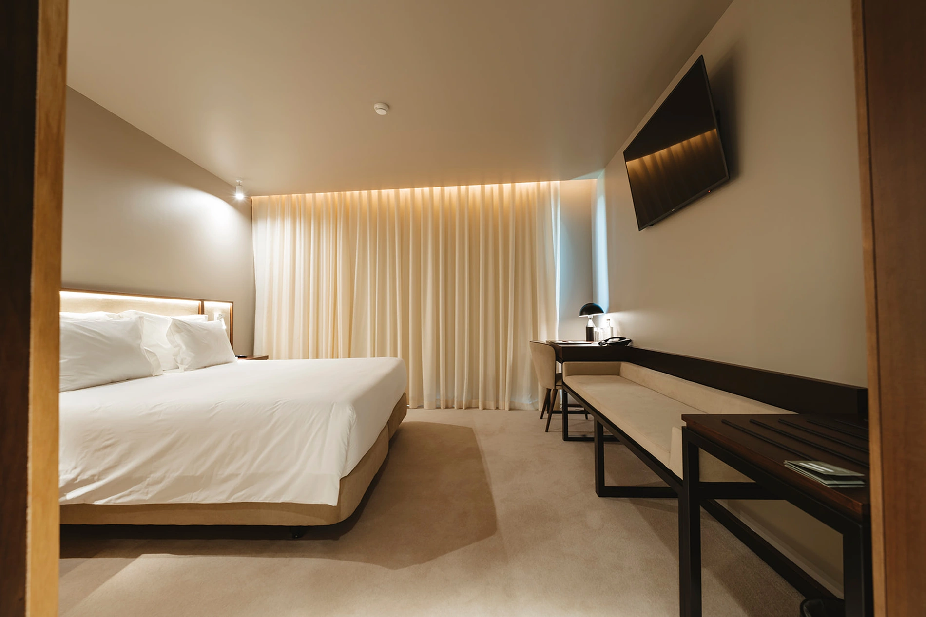 Quarto de hotel moderno com cama, TV, mesa e cortinas elegantes.