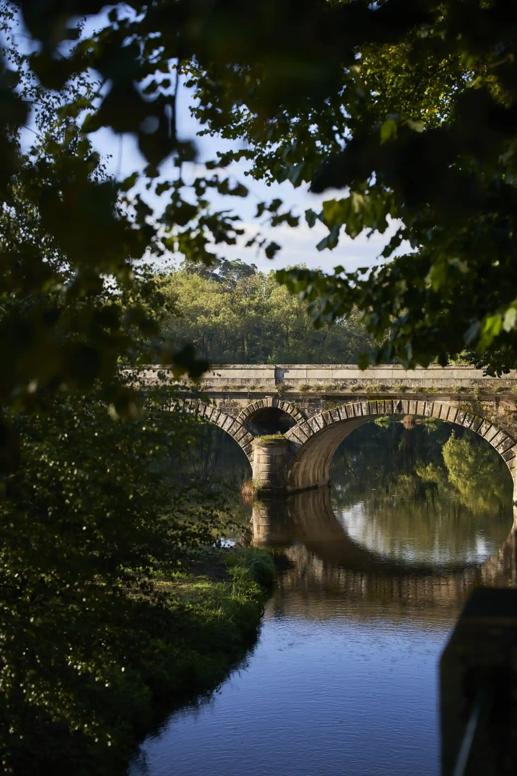 Ponte de pedra sobre rio cercado por &aacute;rvores, refletido na &aacute;gua.