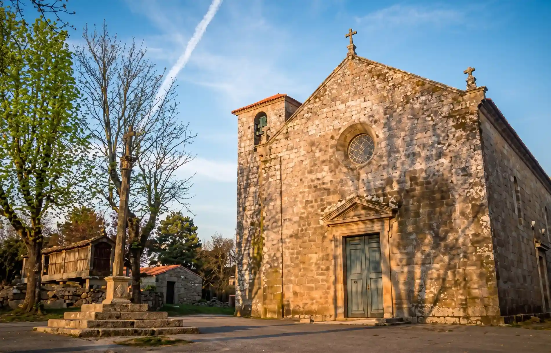 Igreja de pedra ao entardecer, &aacute;rvores ao redor e c&eacute;u claro.