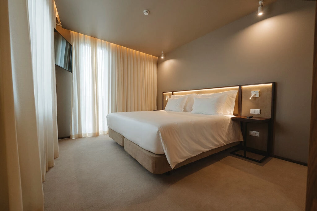 Quarto de hotel minimalista com cama de casal e cortinas brancas.