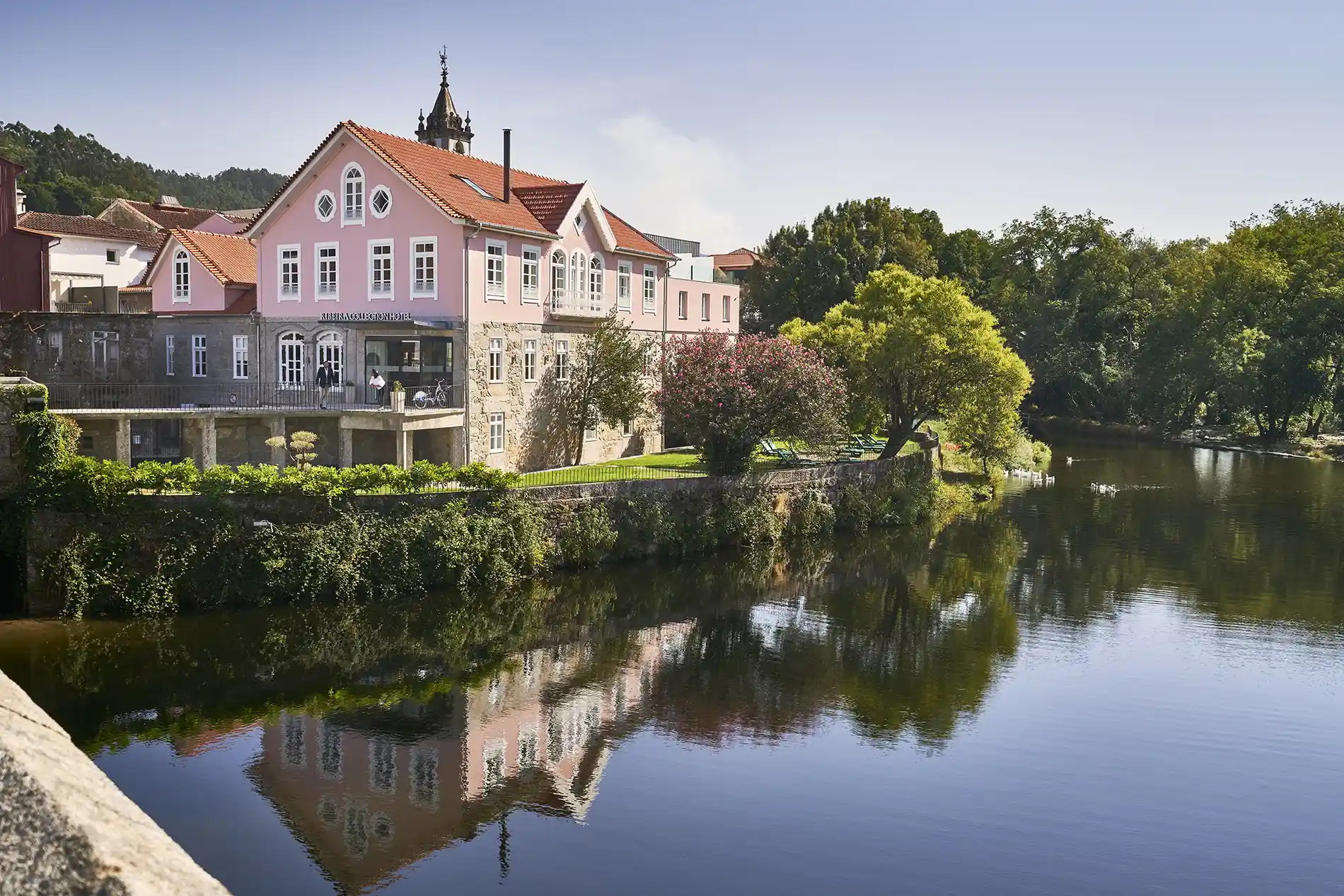 Casa rosa &agrave; beira de um rio com &aacute;rvores e reflexo na &aacute;gua.
