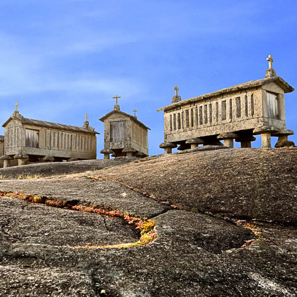 Espigueiros de pedra sobre ch&atilde;o rochoso, com c&eacute;u azul ao fundo.