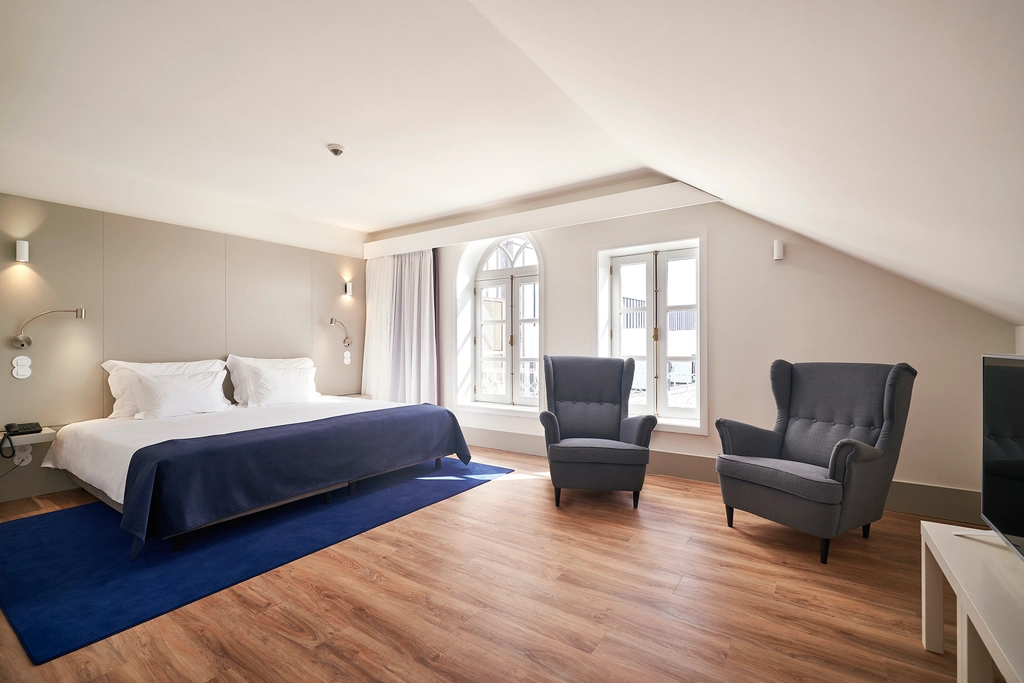 Quarto moderno com cama grande, tapete azul e poltronas perto de janelas.