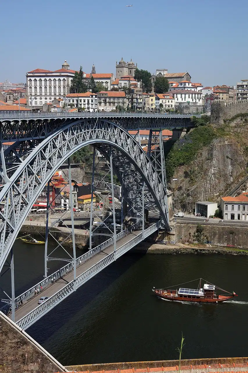 Ponte met&aacute;lica sobre rio, com barco e cidade ao fundo.