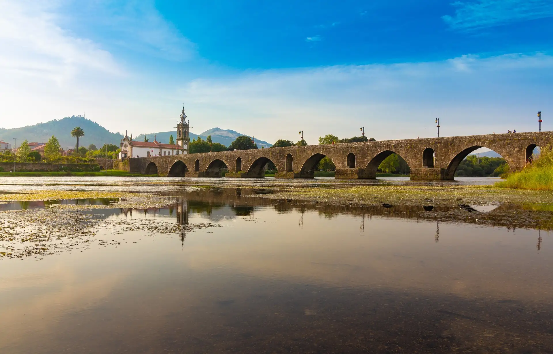 Ponte de pedra antiga sobre rio com igreja ao fundo.