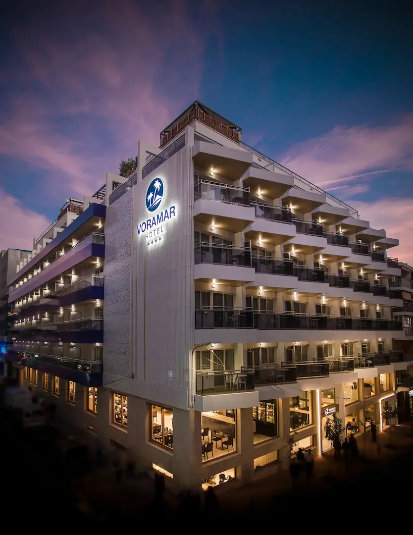 Edificio del hotel VORAMAR con luces y balcones en atardecer, fachada iluminada.