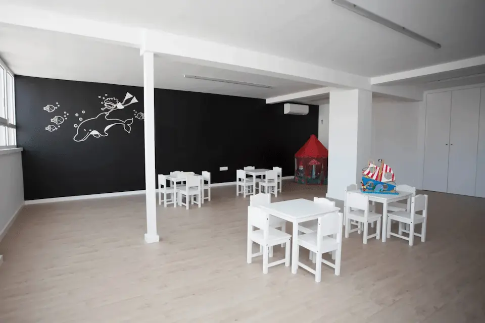 Aula infantil con mesa y sillas blancas, &aacute;rea de juegos y pizarra con dibujos