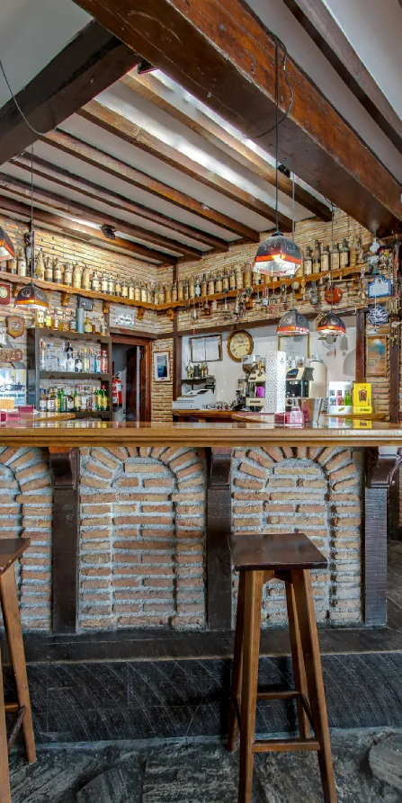 Interior de bar con barra, estantes y luces colgantes bajo vigas de madera.