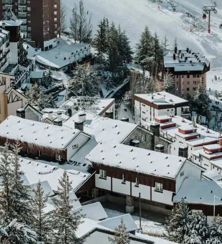 Vista a&eacute;rea de un pueblo nevado entre edificios y pinos, en la monta&ntilde;a.