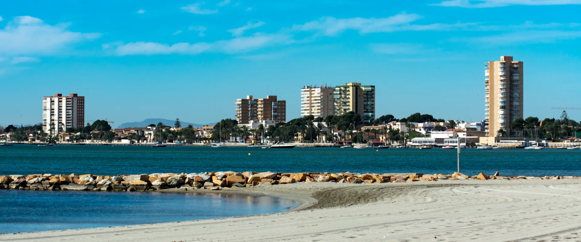Playa de arena con rocas, edificios al fondo y cielo despejado.