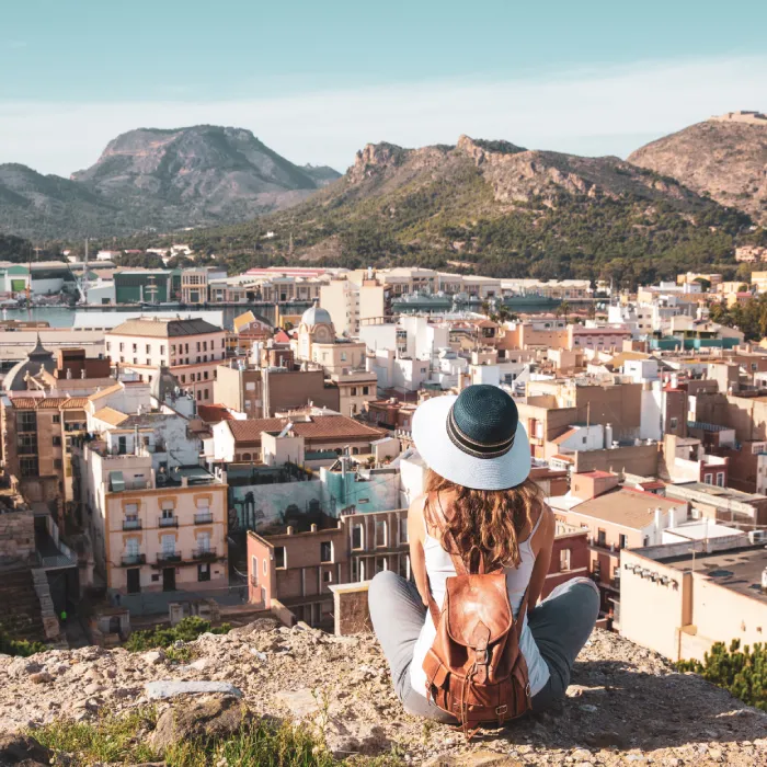 Persona con sombrero y mochila observa una ciudad desde una colina con monta&ntilde;as.