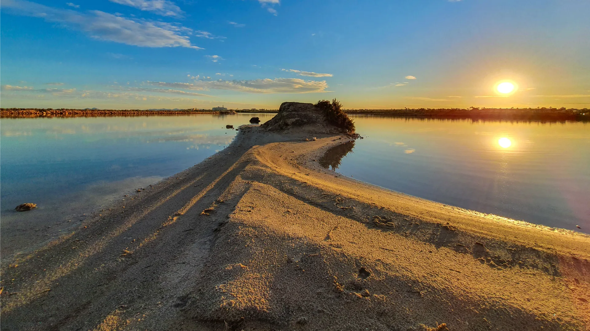 Atardecer reflejado en aguas tranquilas con arena en primer plano.