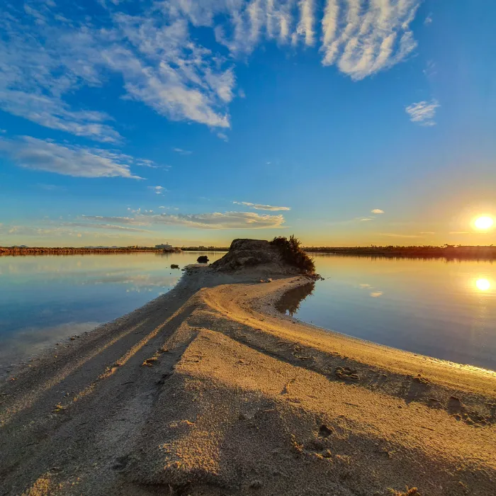 Camino de arena al atardecer sobre un lago con cielo parcialmente nublado.