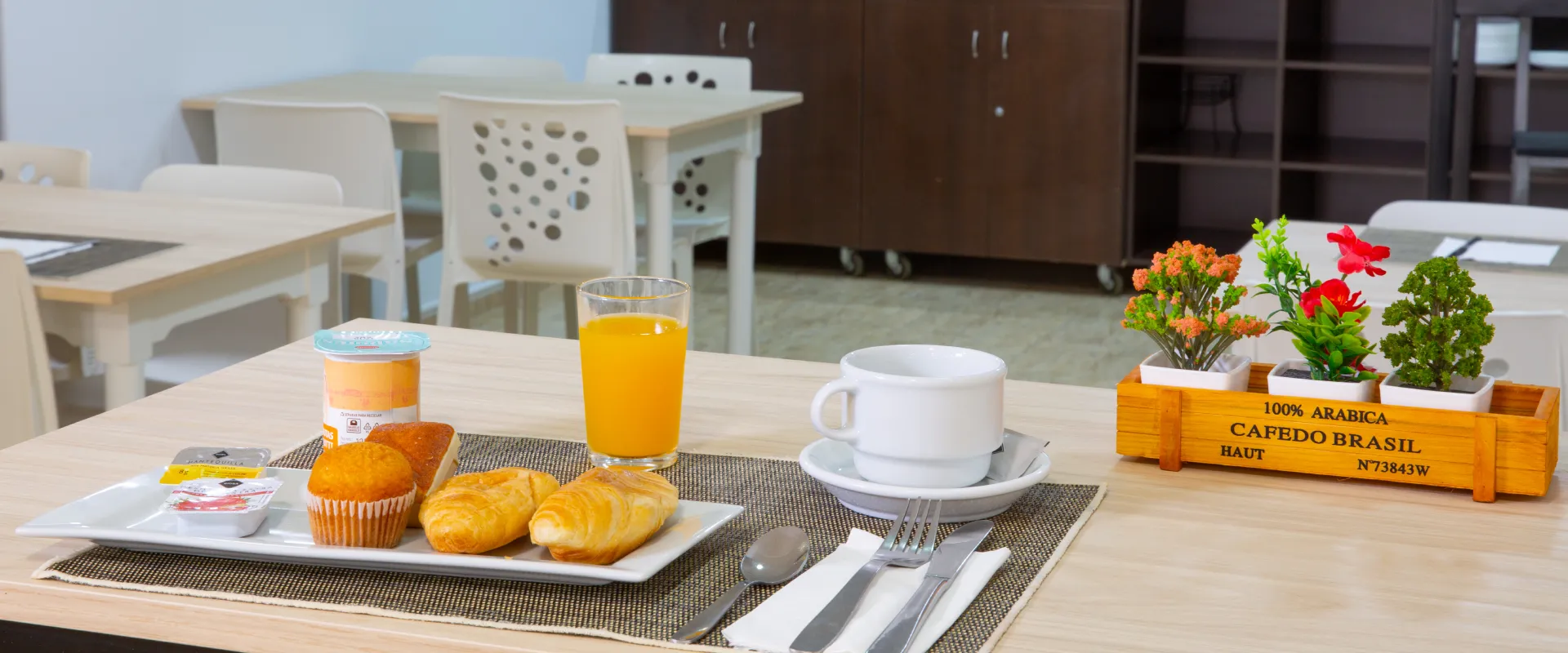 Desayuno con jugo, caf&eacute;, yogur y pasteles sobre una mesa con plantas decorativas.
