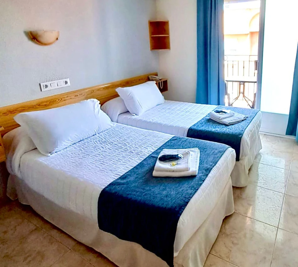 Habitaci&oacute;n con dos camas, s&aacute;banas blancas y mantas azules, junto a ventana