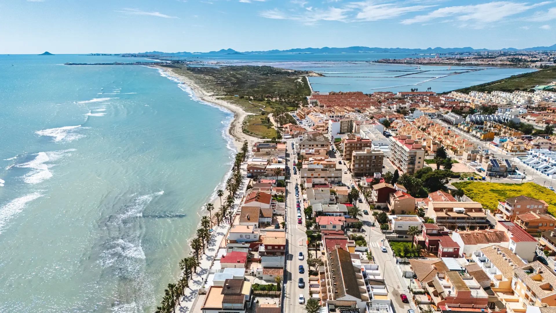 Vista a&eacute;rea de una costa junto a un pueblo con edificios y monta&ntilde;as al fondo.