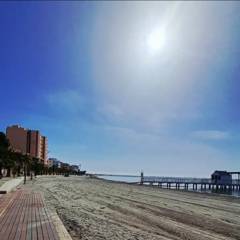 Playa soleada con paseo mar&iacute;timo y muelle en el horizonte.