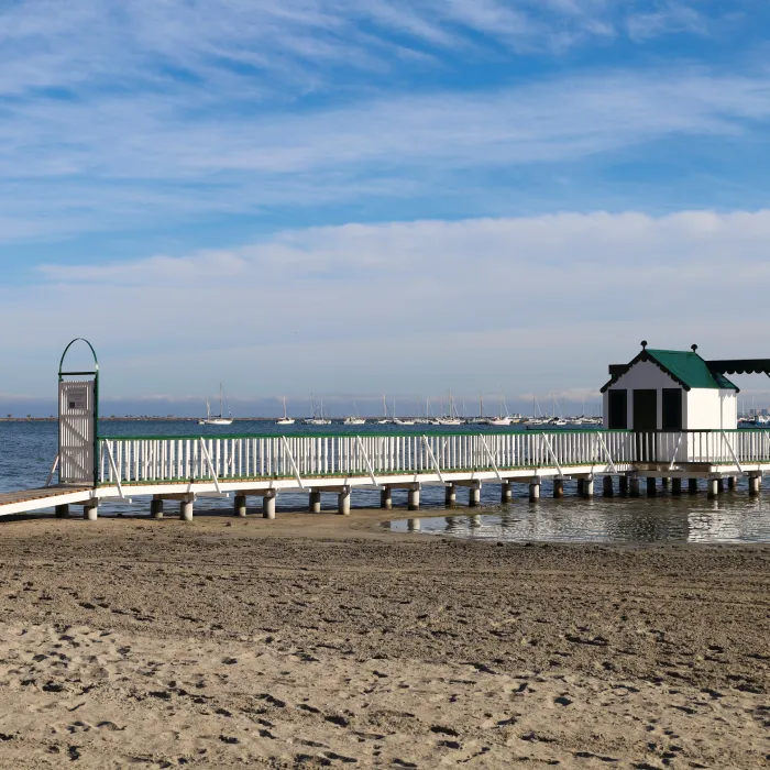 Playa con muelle de madera blanca y caseta, barcos al fondo en el mar.