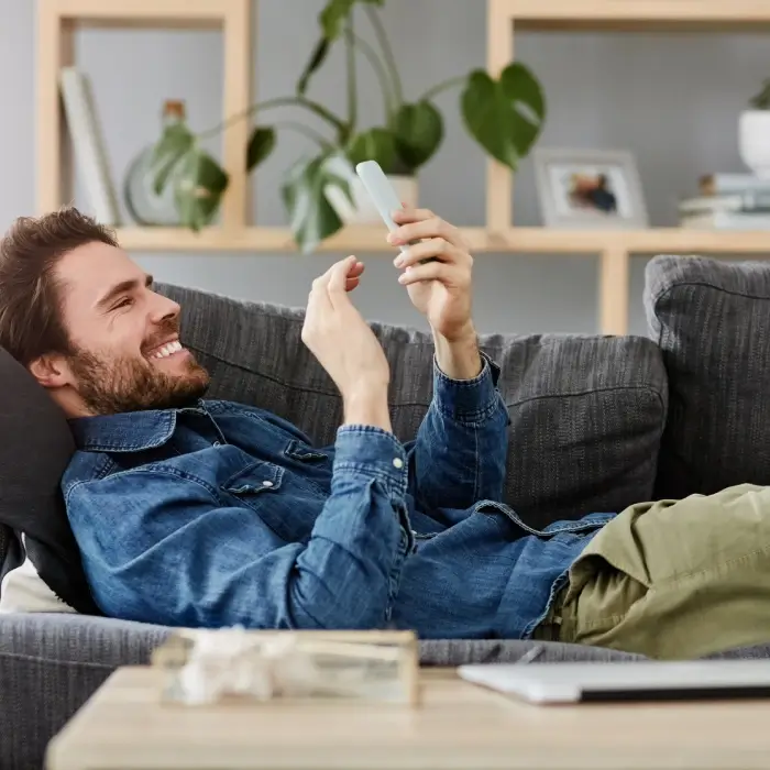 Homme souriant allong&eacute; sur un canap&eacute; regardant son t&eacute;l&eacute;phone.