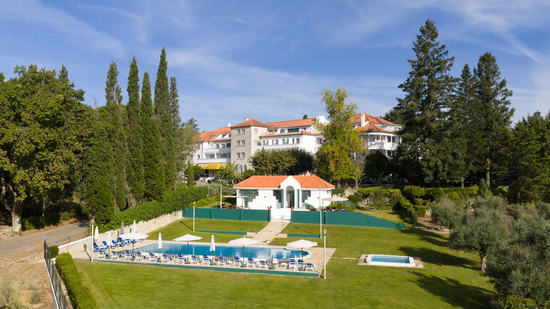 Hotel branco com telhado vermelho, piscina e jardins em dia de sol