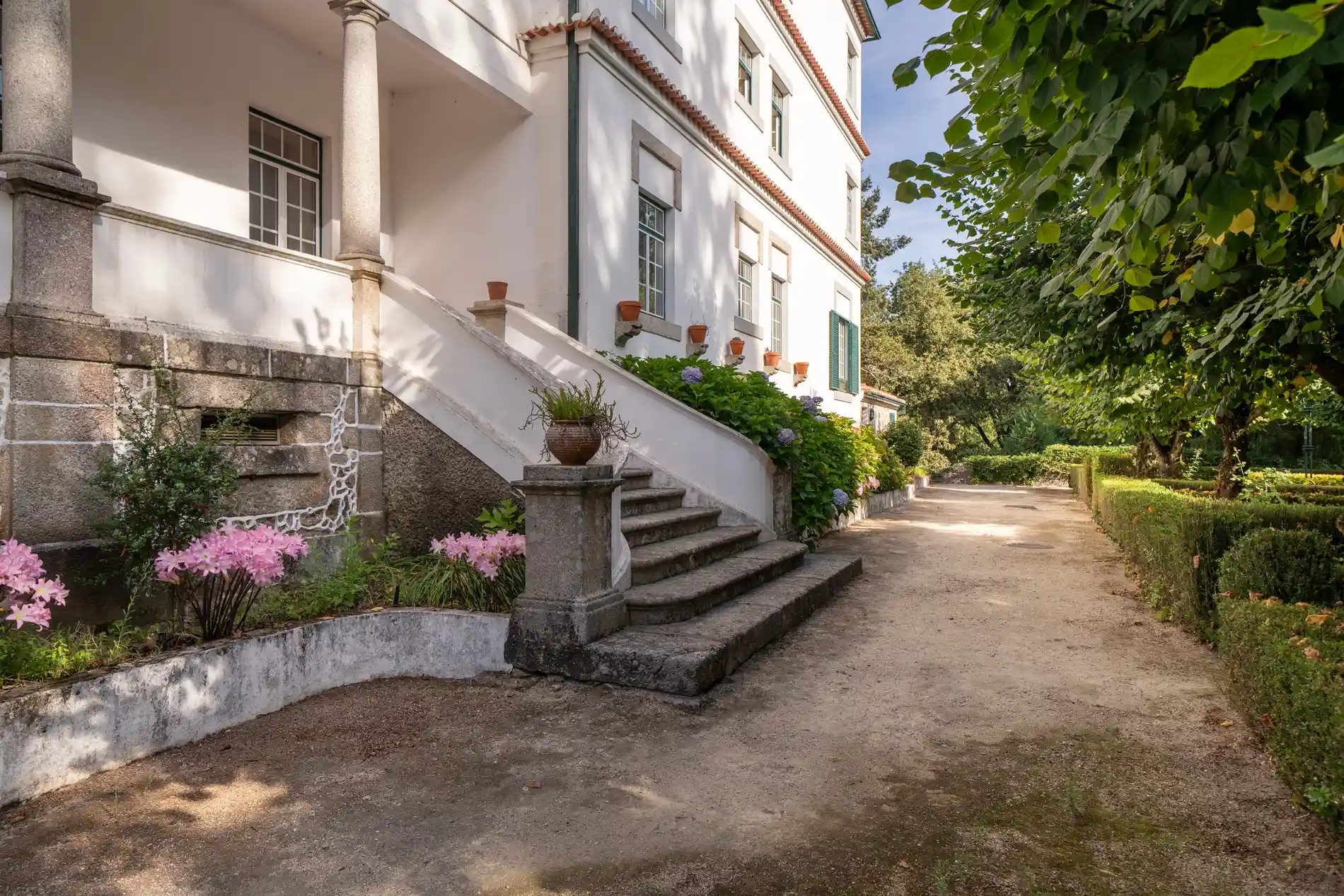 Corredor lateral de prédio branco com escada, vasos e jardim verde florido