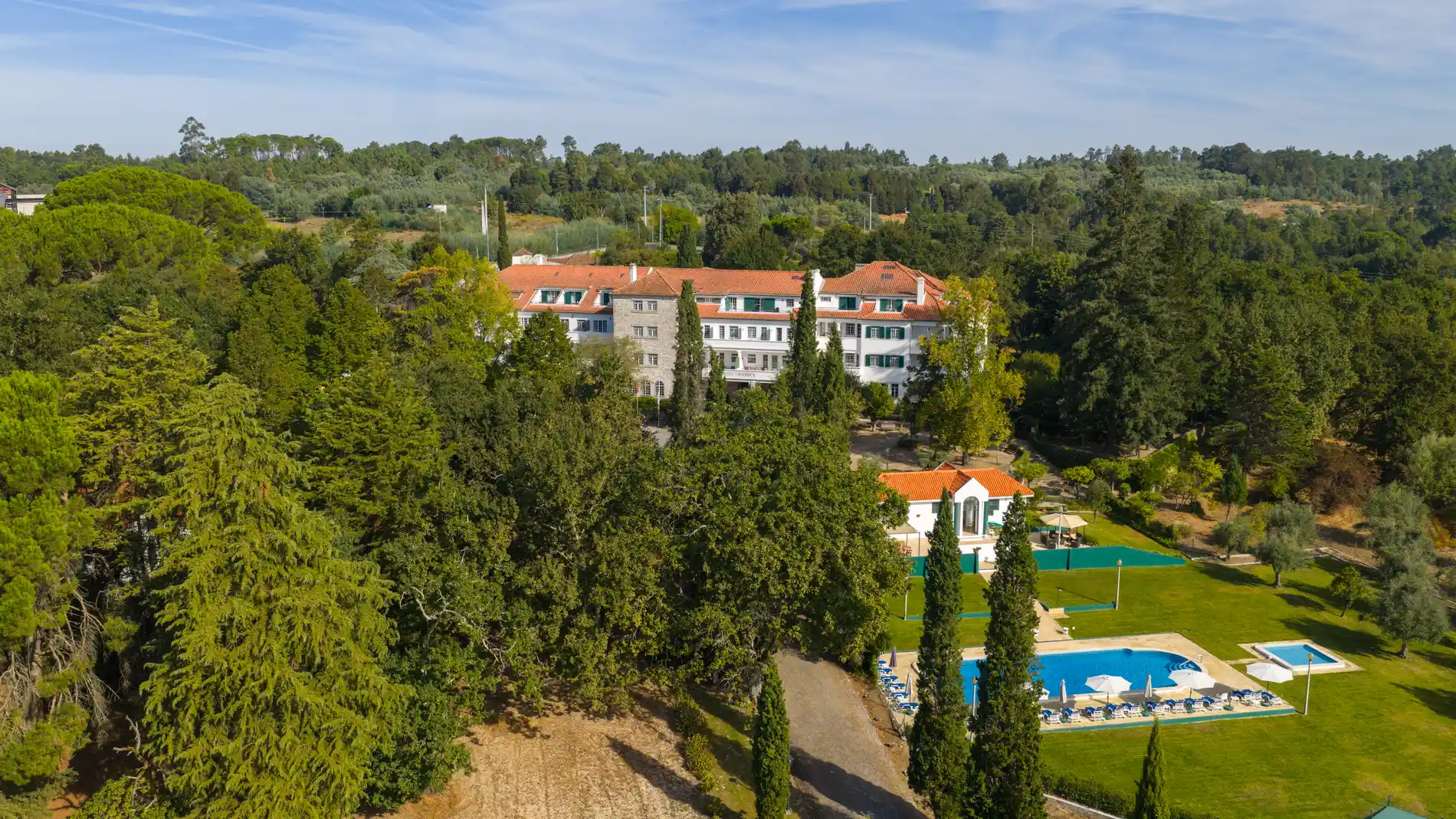 Vista a&eacute;rea de hotel branco com jardim verde, piscina e telhados vermelhos