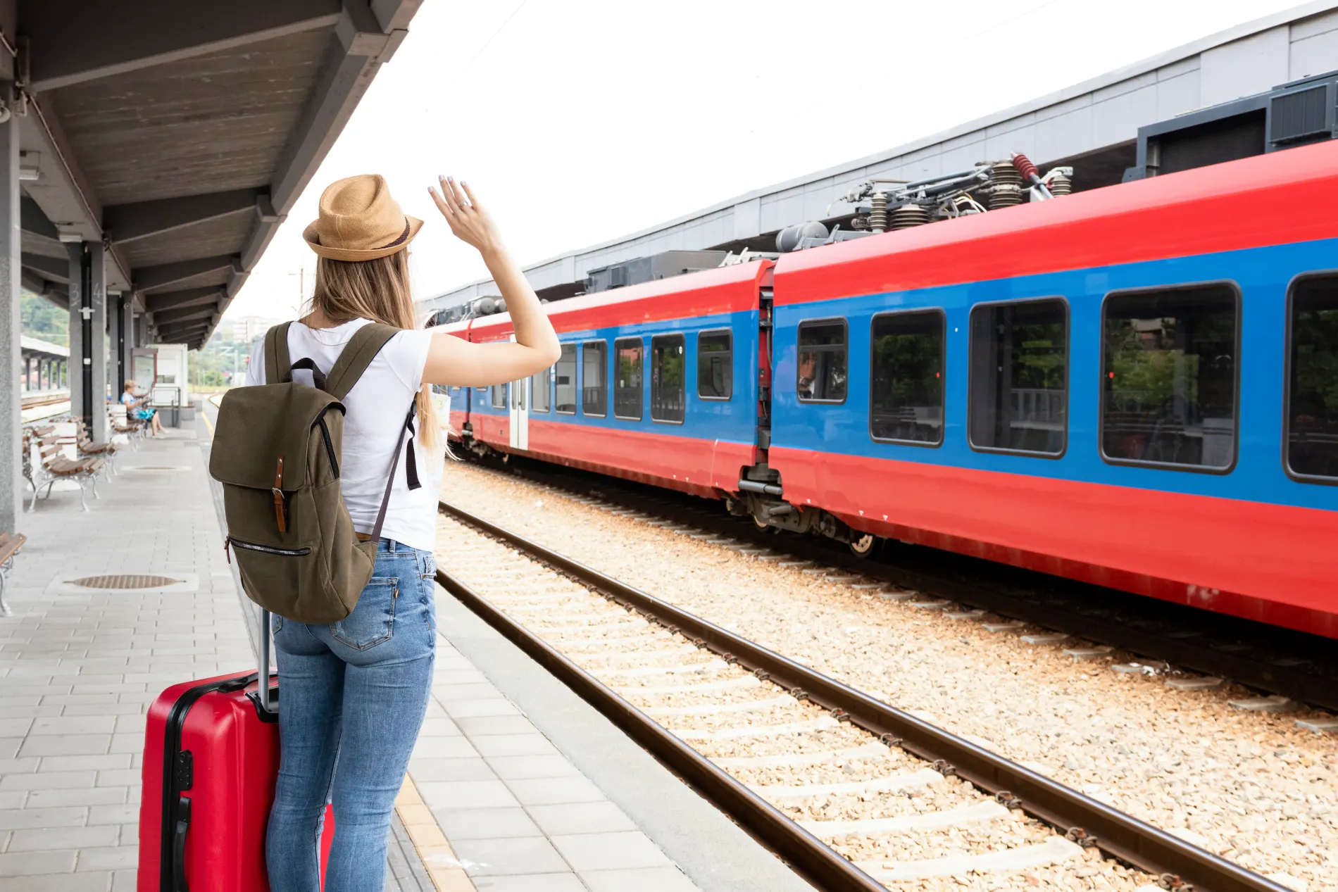 Mulher acenando ao trem azul e vermelho na plataforma, com mala vermelha.