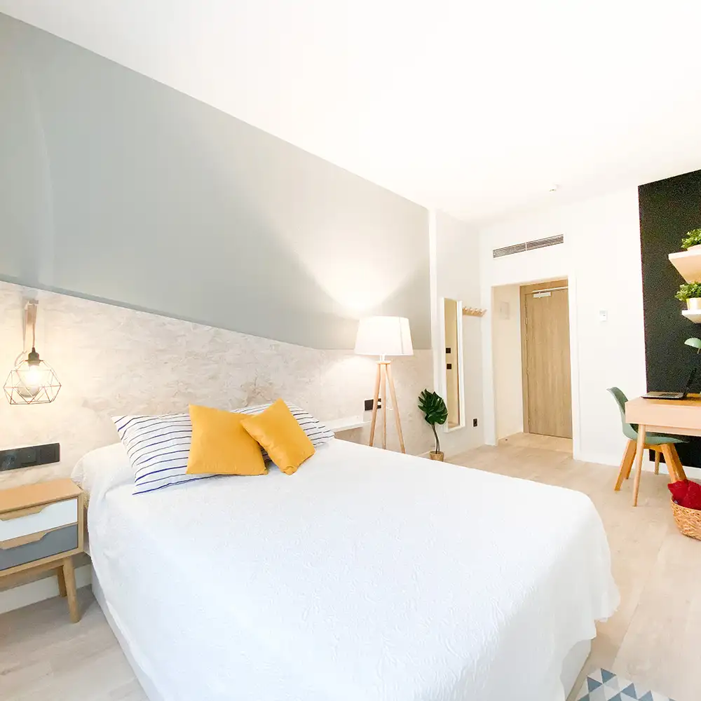 Dormitorio luminoso con cama blanca, almohadas amarillas y mesita con l&aacute;mpara
