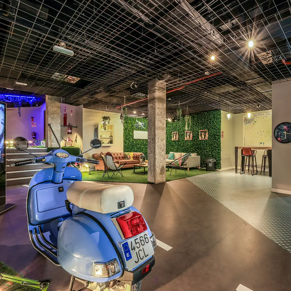 Interior con pared verde, sof&aacute;s y una Vespa azul en primer plano bajo rejilla de techo